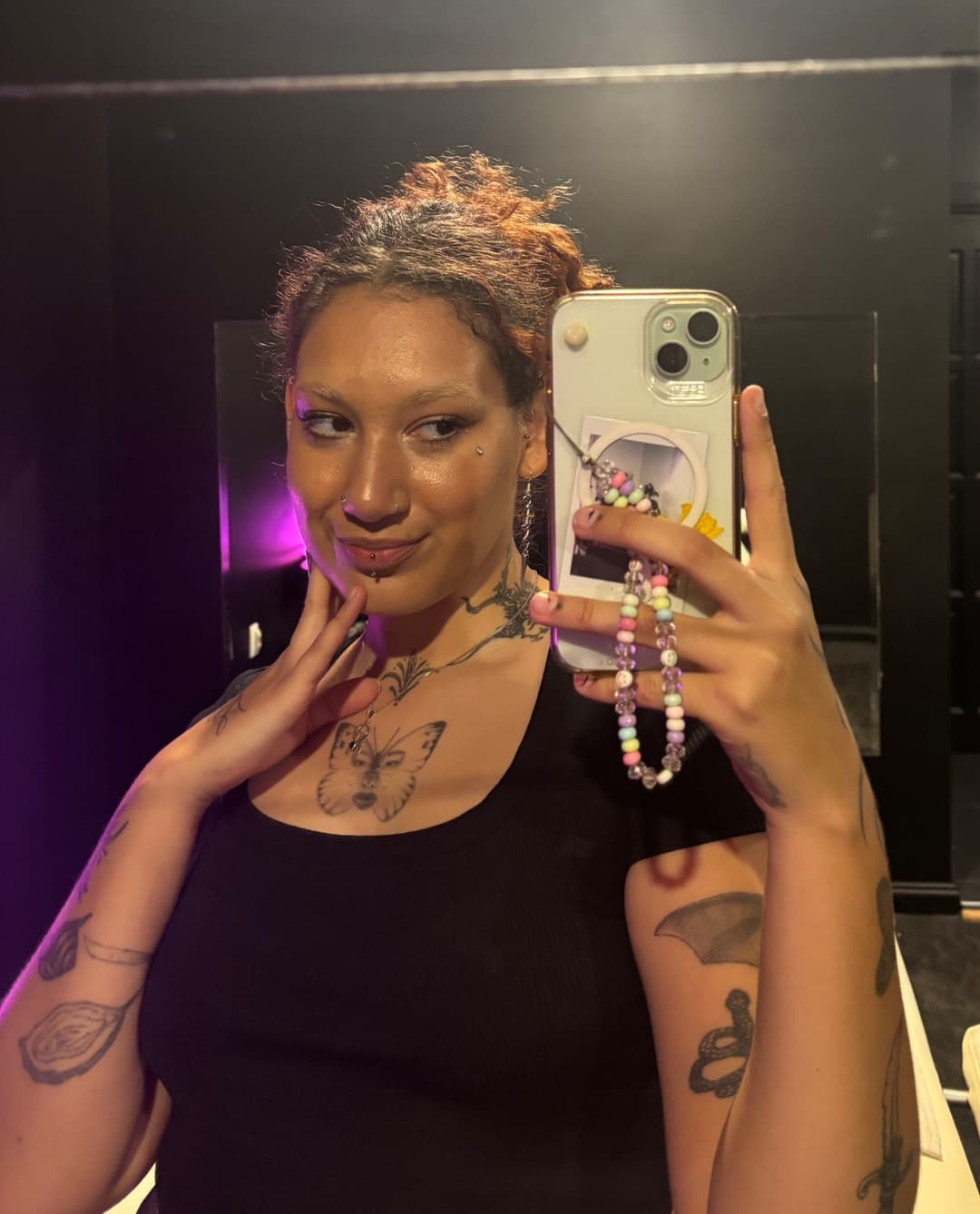 A person with curly hair tied back takes a mirror selfie in a dimly lit room, wearing a black top. An independent masseur, they have tattoos—including a butterfly on their chest—and hold a phone with a colourful beaded charm.