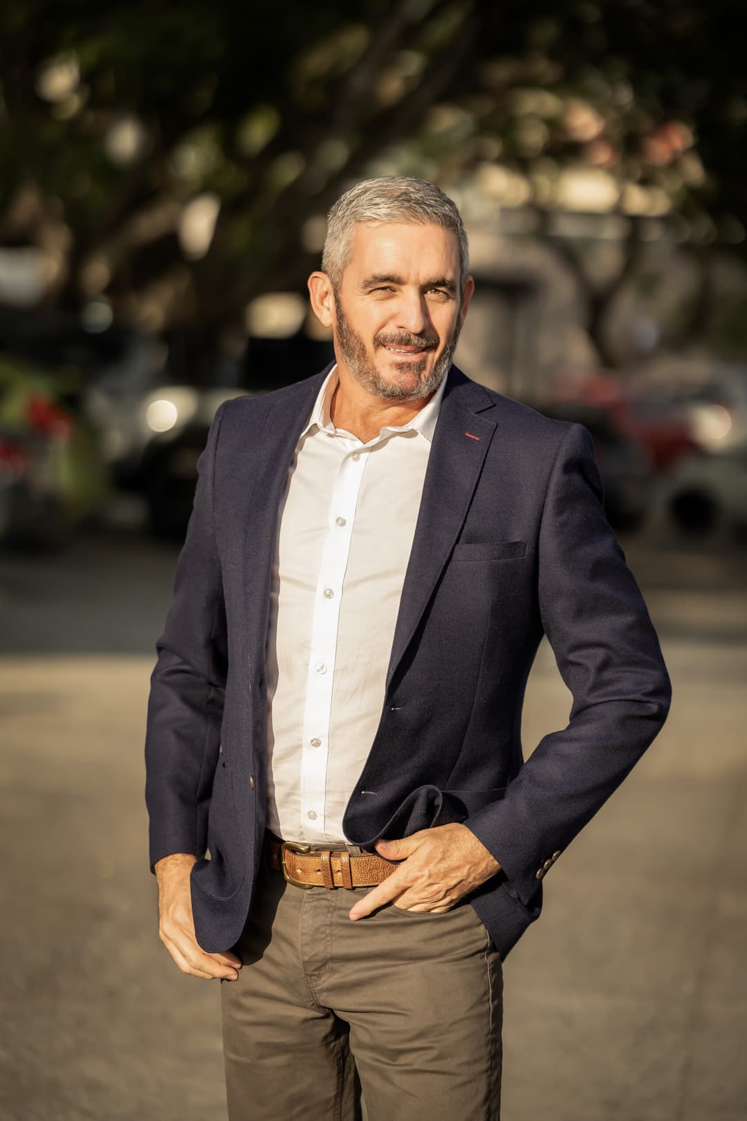 A man with short grey hair and a beard, wearing a navy blazer, white shirt, and brown belt, stands outdoors on a sunny day, smiling with one hand on his hip. The backdrop hints at relaxation found in sensual massage getaways.