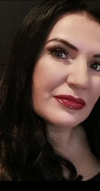 A woman with long dark hair, bold eyebrows, dark eye make-up, and glossy red lipstick looks towards the camera with a slight smile, capturing the allure of an independent masseuse against a dark background.