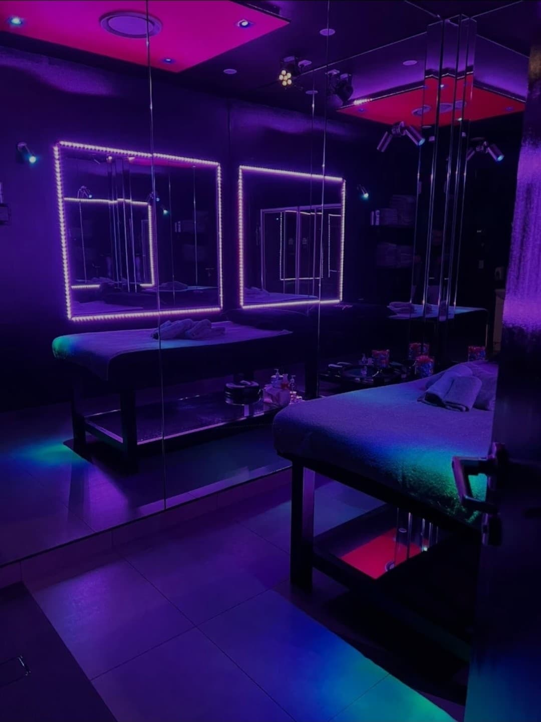 A dimly lit spa room at Mistiques Newtown Massage Salon features two massage tables, square mirrors outlined with purple LED lights, and colourful ambient lighting in shades of pink, purple, and blue reflecting on the floor and ceiling.