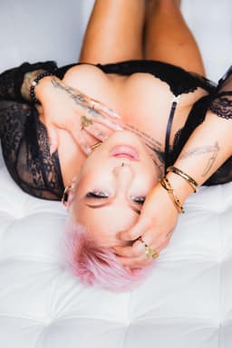 A person with short pink hair and tattoos lies on a white surface in black lace lingerie. Their hand rests on their face, adorned with rings and bracelets, exuding confidence—like an independent masseur offering tantric massage Australia.
