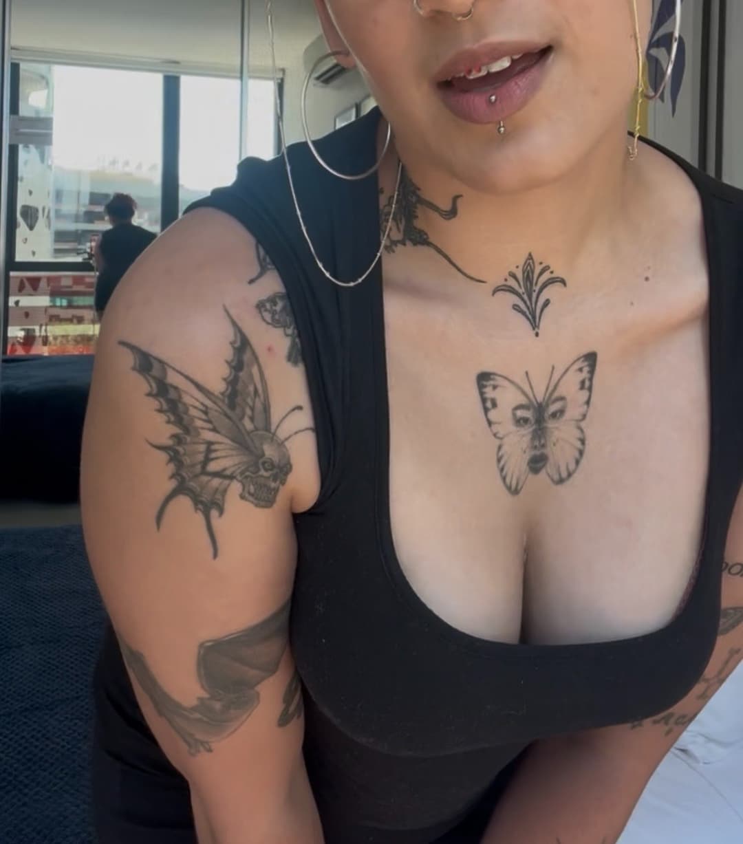 A person with short hair and facial piercings is leaning forward, wearing a black sleeveless top. Visible tattoos include a butterfly and a skull-winged insect on their shoulder, and a butterfly on their chest.