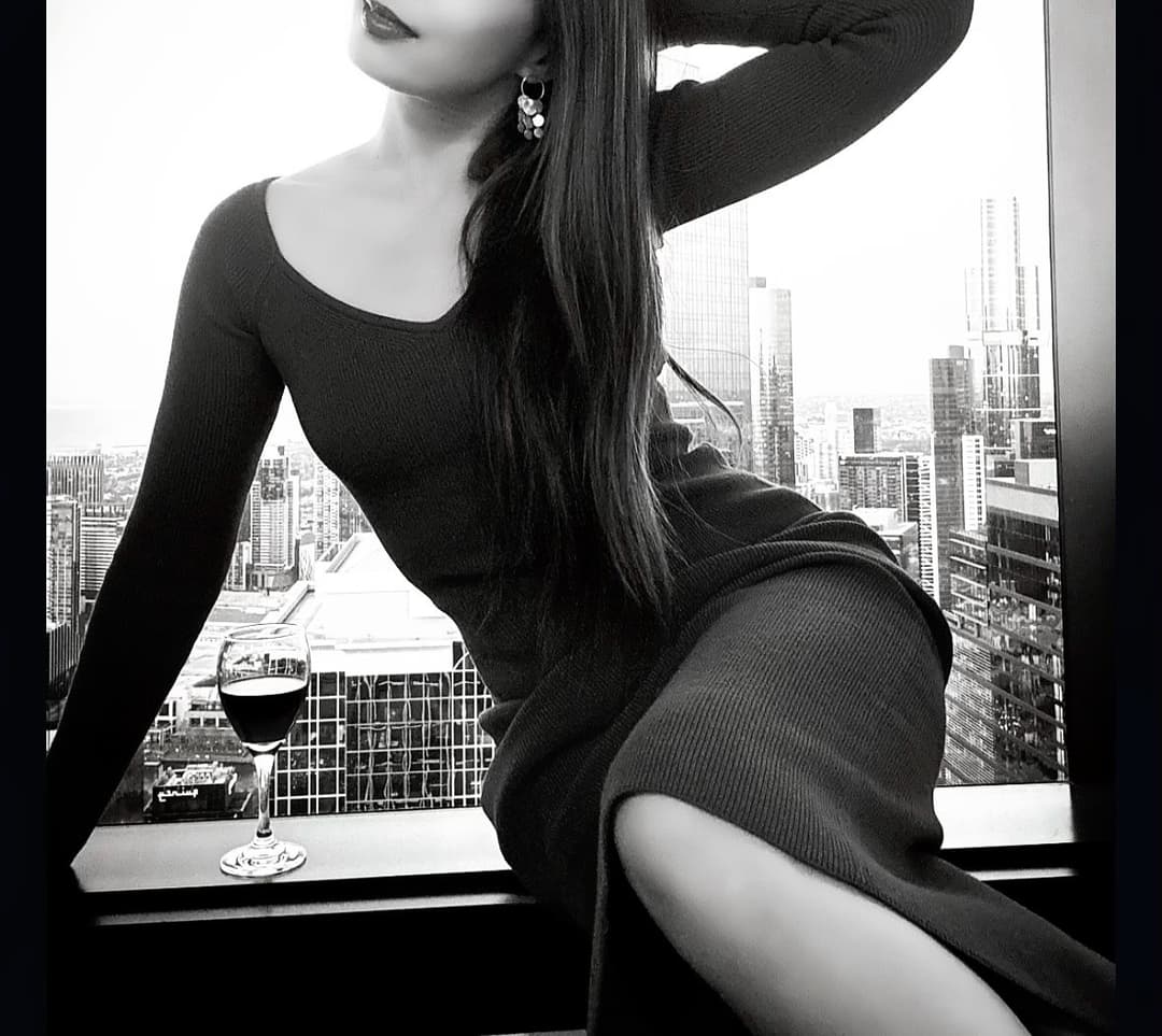 Tantric practitioner Eden sitting on a window sill with a glass of wine, overlooking the Melbourne city skyline during an intimate, sensual moment