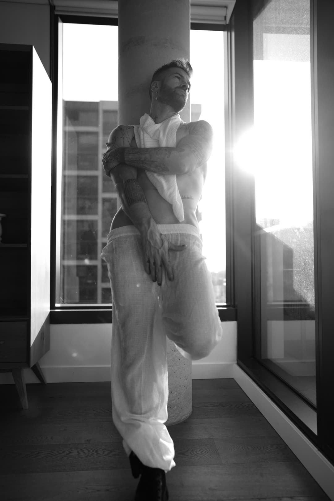 Lucian Blake posing in sheer white clothing by a window in black and white, featured profile image for professional sensual massage services.