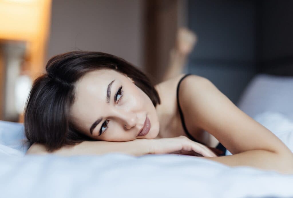 A woman with short dark hair lies on her stomach on a bed in an inviting incall setting, resting her head on her arms and gazing thoughtfully to the side. She is wearing a black strappy top and the background is softly blurred.